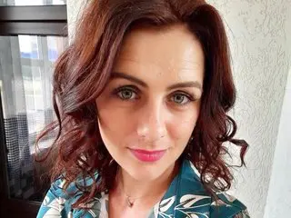 Portrait of a smiling woman with wavy red hair wearing a teal floral patterned blouse in a bright indoor setting.