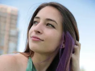 Magnoliajoy looks at the camera with a soft smile, her long brown hair falling over her shoulders against a bright, outdoor background.