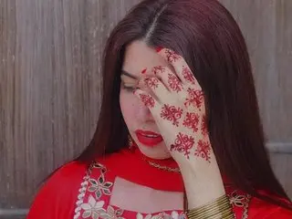Mahrukh has long dark hair and red lips, posing with her henna-decorated hand over her eyes while wearing a red outfit.