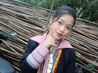 Maimai555 poses with a hand to her chin, wearing a black top with pink and white patterned trim, her dark hair pulled back in a simple setting.