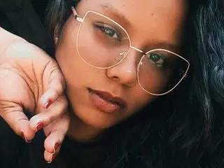 Mairaasm looks into the camera with a close-up view of her face, wearing gold-rimmed glasses and letting her dark hair fall around her face.