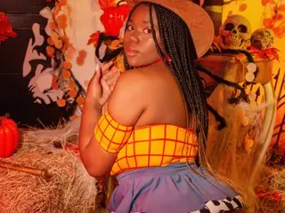 MakenaGolden poses with her back turned, showing her bare shoulders and long dark braids against a Halloween-themed background.