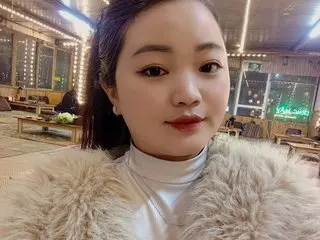 Young woman in a white turtleneck and fluffy faux fur coat smiling in a cozy, lit restaurant setting.