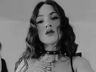 ManuEvans poses with her wavy hair down, wearing a black choker and a black bra or top against a plain background.