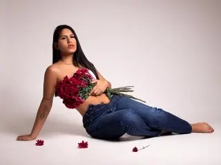 Maragirl poses with long dark hair, wearing a red floral bustier and blue jeans while holding a bouquet of red roses against a plain white background.