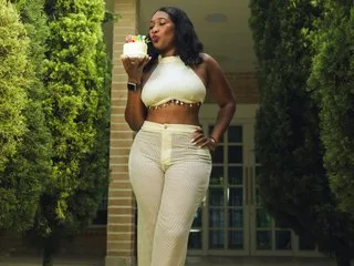 MarahJones wears a white halter top and matching pants while eating a small cake outdoors near green bushes.