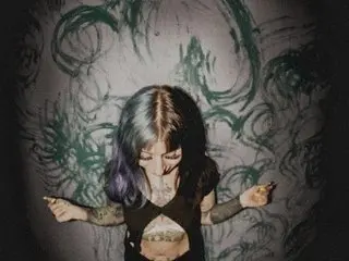 MarcelineWaters poses with her long blue and purple hair, wearing a black bra that shows off her breasts and stomach against a dark, swirly background.