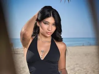 Marianamoon19 poses with her hands in her dark hair, wearing a black low-cut bodysuit against a beach background.
