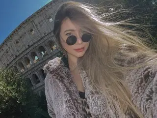 Marieleclerc with long blonde hair and sunglasses posing in a fuzzy grey coat in front of the Colosseum.