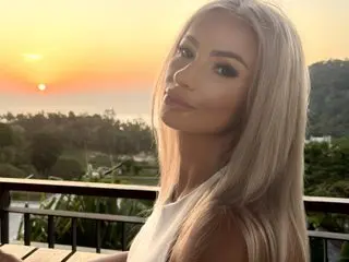 Blonde woman in a white top enjoying a golden sunset view from a tropical balcony.