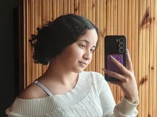 Marilu_Malibu poses with a soft smile, wearing a white off-the-shoulder sweater and holding a phone up to her face in a close-up shot.