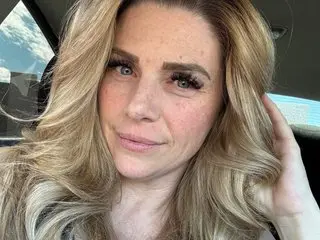 Smiling blonde woman with freckles and long wavy hair sitting in a car on a bright day.