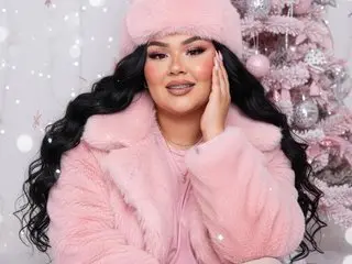 MarinaBusty poses with long wavy black hair, wearing a pink furry coat and hat against a soft pink background with light sparkles.
