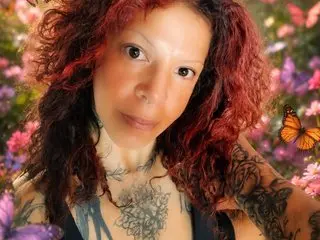 Mariposatatto has long red hair and visible neck and chest tattoos while posing in a garden setting.