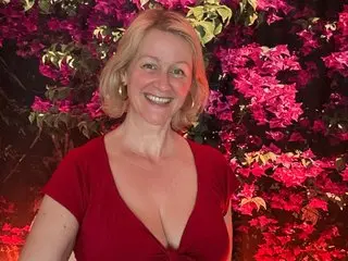 MarleneBloem smiles while posing outdoors against a background of bright pink flowers, wearing a low-cut red top that shows her cleavage.