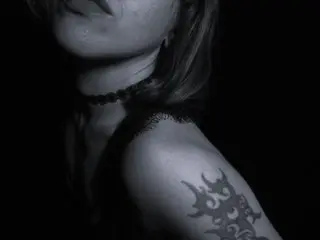 MartaShadow poses in a dark setting, showing her neck, shoulder, and a large black tattoo on her arm.