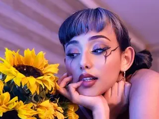 Young woman with artistic eye makeup posing dreamily with bright yellow sunflowers in soft purple lighting.