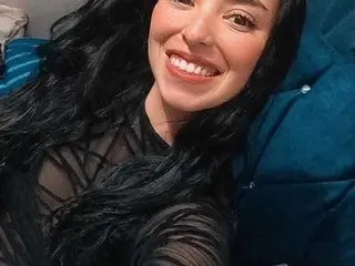 MartinaReymond smiling with long wavy black hair while wearing a black sheer mesh top.