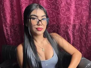 MarySokova poses with long black hair and glasses, wearing a light grey tank top against a pink patterned background.