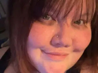 MaryWett smiles close-up, showing her face and light brown hair with bangs in a dim indoor setting.
