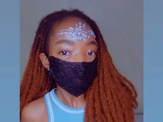 MaskedEbonyPrincess poses with long brown dreadlocks and white glittery face paint or jewels on her forehead, wearing a black lace mask and a light blue top.