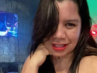 MatureAnika smiles while posing with her hand against her neck, showing her long dark hair and bare shoulders.