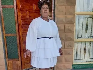 MatureTemptress00 — Stylish woman in a white tiered dress and black belt standing outdoors by a wooden door.