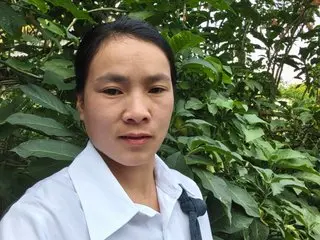 May_Vietnamese poses outdoors against green leaves, wearing a white collared shirt with her dark hair pulled back.