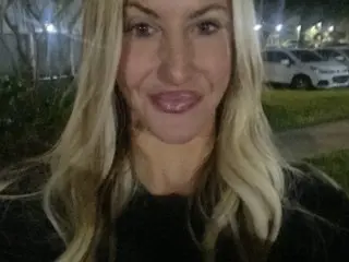 Smiling blonde woman in a black top outdoors at night near parked cars.