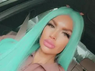 Woman with long mint green hair and pink makeup sitting in a car wearing a mauve blouse.