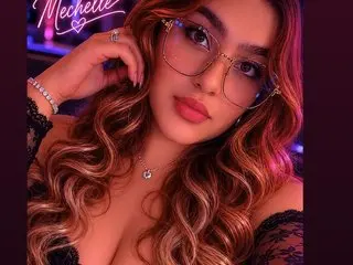 MechelleLyonn poses with long wavy hair, wearing a black lace top and glasses, looking directly at the camera.