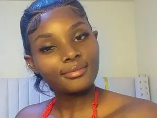 Smiling young Black woman with sleek hair and red beaded necklace in a bright bedroom setting.