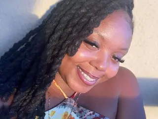 MeetGraceGold smiles while posing close-up, showing her long dreadlocks and wearing a floral top against a plain wall.