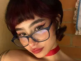 Young woman with short dark hair and glasses wearing a red choker in a warm indoor setting.