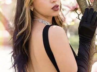 MeganLewits poses outdoors in a black dress and long black gloves, leaning against a tree branch.