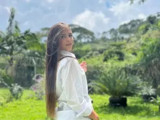 MeganVellvet poses outdoors with long brown hair, wearing a white button-down shirt against a lush green garden background.