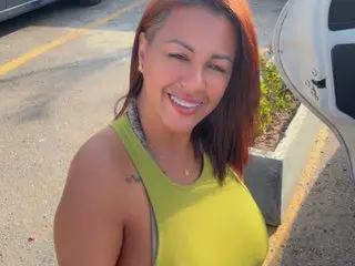 Megangreyxx smiles while wearing a yellow tank top, showing her long reddish-brown hair and a small tattoo on her shoulder outdoors.