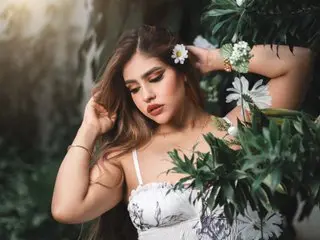 MelaniIeRioss poses with her long brown hair, wearing a white lace bra or camisole against a lush, leafy green background.