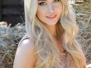 MelanieSpaces smiles with her long blonde hair down, wearing a sparkly pink top while posing against a hay background.