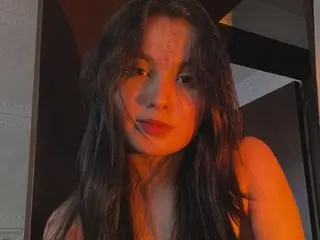 Melanieeev poses with long dark hair, looking directly at the camera with a soft expression in a dimly lit indoor setting.