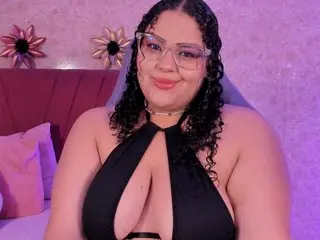 Melannie_Carter poses in a black halter top, showing her cleavage and curly dark hair against a soft pink background.