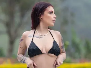 MelannyLuuv poses with her hands near her waist, wearing a black string bikini top and showing her tattoos against a soft, natural outdoor background.