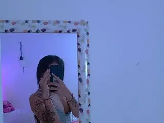 MeliMuller poses for a mirror selfie, showing her face, hair, and chest while holding a phone, set against a plain blue wall.