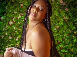 MelisaCarusso24 poses with long braids and hoop earrings against a green leafy background, wearing a white top.