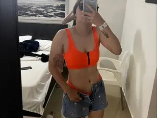 Melissa0506 poses in an orange bra and denim shorts, showing her midriff and cleavage while standing in a room with a bed.