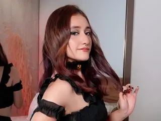 MelissaBlooms poses with her long brown hair and red lipstick, wearing a black ruffled choker and a black top against a soft pink background.