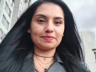 Melissa_Rhodes smiles for a selfie outdoors with her long black hair flowing over her shoulders while wearing a grey shirt and black necklaces.