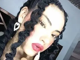 Glamorous woman with long curly black hair, heart earrings, and red lipstick posing in a modern setting.
