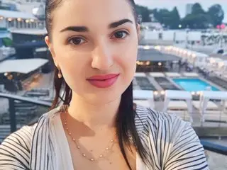 Meri_Gold poses with a direct gaze, wearing a striped top and showing her dark hair and bare shoulders against a bright outdoor background.