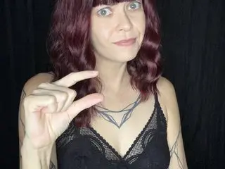 MerlotMistress poses in a black lace bra, looking at the camera with her wavy red hair framing her face against a dark background.